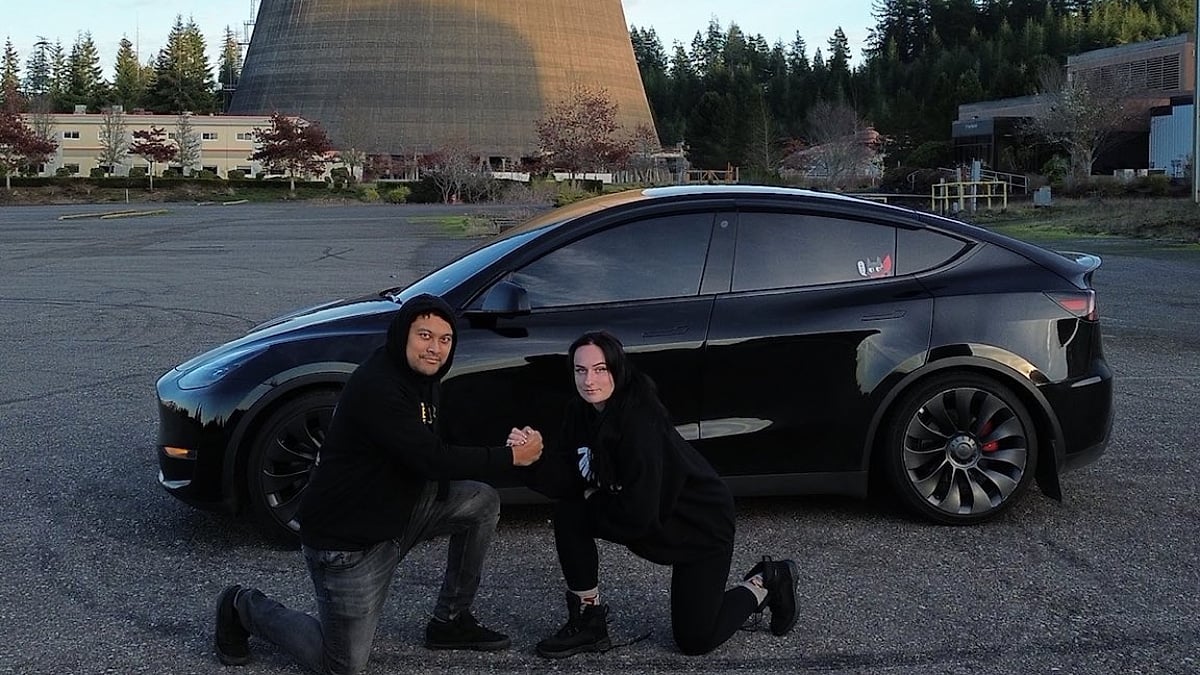 Eddi and Suzie posing beside their black Model Y in a parking lot near an industrial cooling tower.